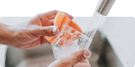 Persona lavando un vaso transparente con una esponja bajo el chorro de agua del grifo.

